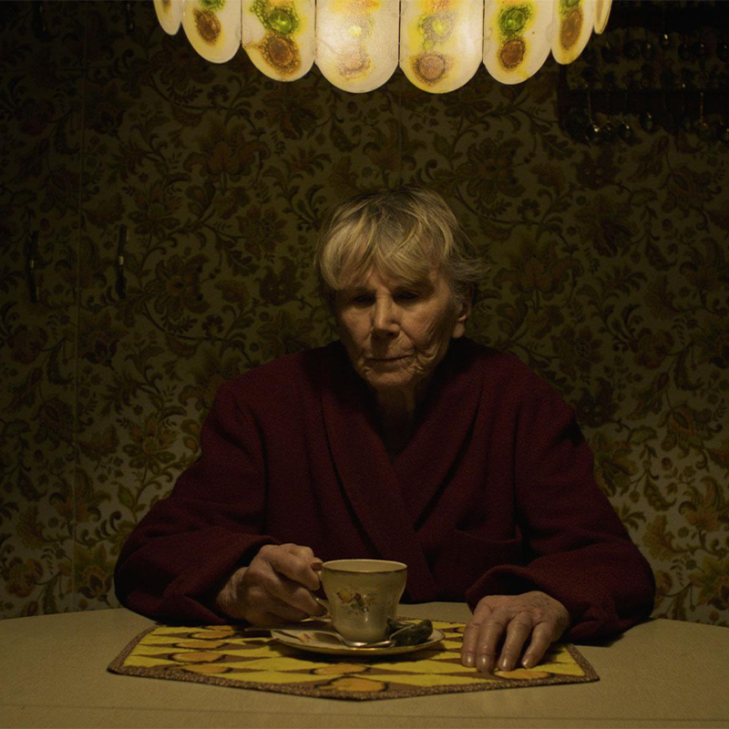 A photo of an older woman sitting alone, in a dimly lit kitchen, at a kitchen table with a tea cup in her hand
