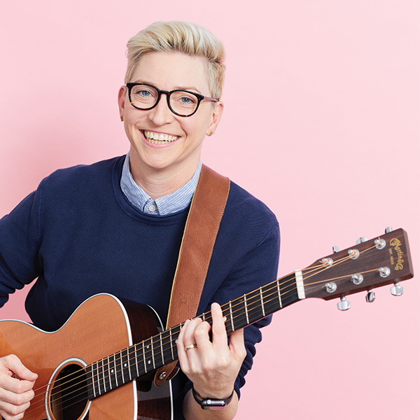 An image of Kristin Key, short blonde hair, thick black glasses playing a guitar, smiling